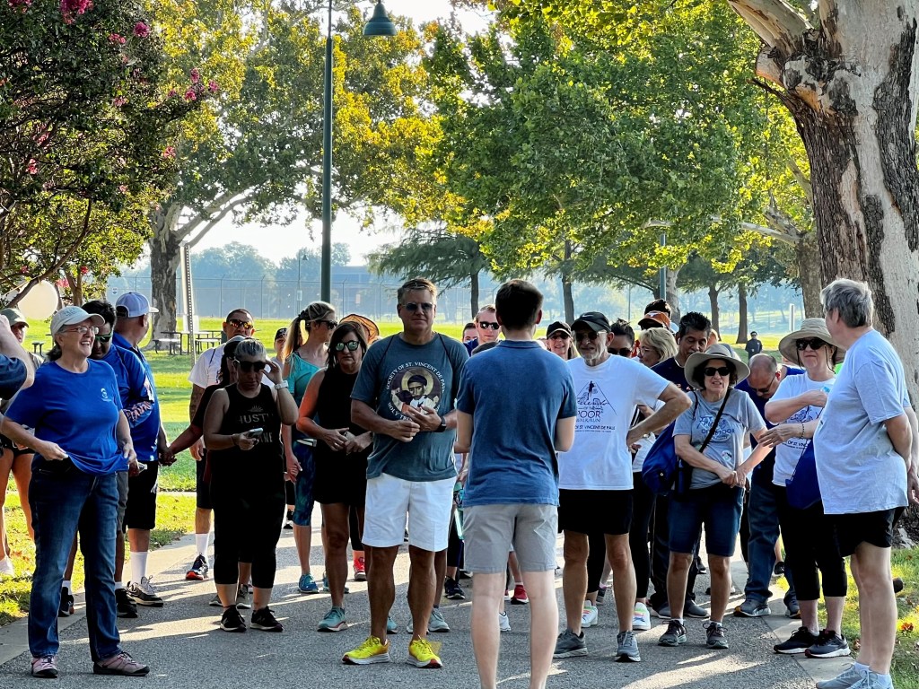 A group of people gather in a park, likely for a walk or event. The group is diverse and varied, with multiple individuals...