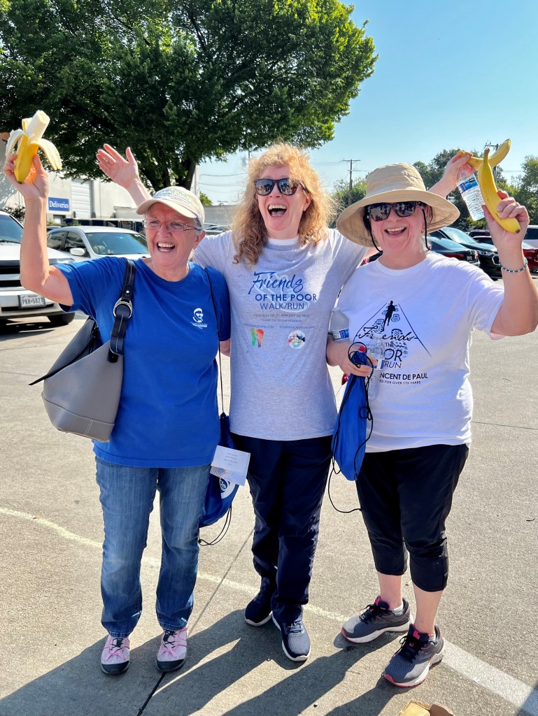 Three women standing side by side, smiling, holding up banana peels dressed in warm weather clothing.