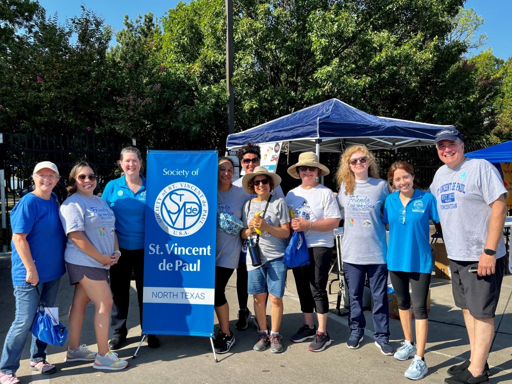 A group of people standing next to a blue banner and a canopy tent, likely at an outdoor event or festival.